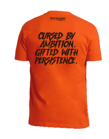 Orange Shirt with Text "Cursed by Ambition. Gifted With Persistence"